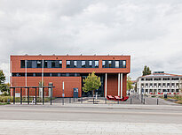 The photo shows two buildings on the campus of Landshut UAS, one has a brick red color, the other is white. A bus stop is in front of the red building. Red benches are scattered around the campus.