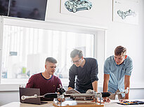 Three students discuss their assignment in a bright room. In front of them sits a model on the desk.  On the wall behind them are two pictures of car models. 