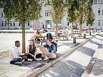 A group of 3 students chat outside on the campus of Landshut UAS, in the background students are sitting on benches.