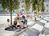 A group of 3 students chat outside on the campus of Landshut UAS, in the background students are sitting on benches.