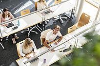Vogelperspektive auf Studierende, die in der Bibliothek lernen, einer davon schaut lächelnd in die Kamera hinauf