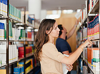A woman uses her hand to look for a book in the library, in the blurry background is a man taking a book from a high shelf.