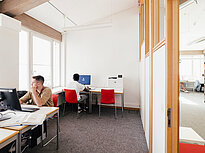 Photo shows two students, each sitting at their own computer in a PC room. The room is bright and has computers as well as a printer. One of the two students is looking outside the window.