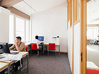 Photo shows two students, each sitting at their own computer in a PC room. The room is bright and has computers as well as a printer. One of the two students is looking outside the window.