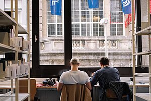 Students in the Library @Anna Tiessen Two students sit next to each other in front of a big window in the library. Outside the window are three flags each with a letter from "F", "A", and "U" from left to right.