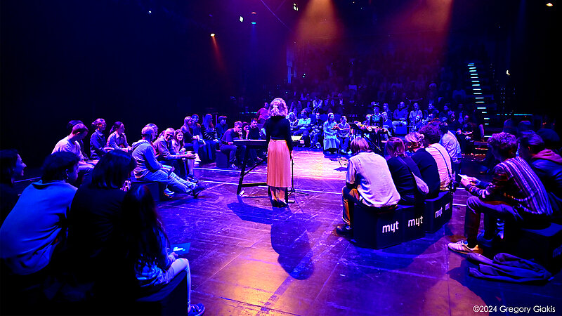 @ HMTM In dunklem, buntem Licht sitzen Menschen in einem großen Saal, in der Mitte steht eine Frau an einem Keyboard