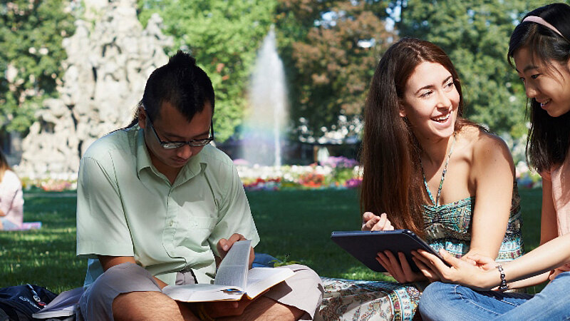 Students Studying in the Park @ FAU Three students with diverse backgrounds sit on the grass reading and talking together. In the background is a fountain and flower garden