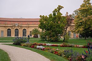 Erlangen Orangery @Anna Tiessen In the foreground is a path, on the side are colorful flower bushes and green grass. The Erlangen Orangery lies in the background, partly hidden behind trees.