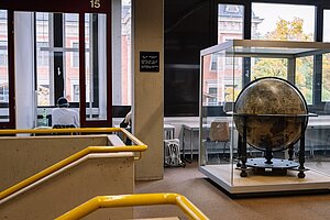 Erlangen Library with a Globe @Anna Tiessen The photo shows a part of the stairs on the upper floor with bright yellow handrails, near the stairs is a big globe in a glass case. Nearby students are studying.