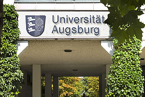 @ Uni Augsburg Ein Außenflur an der Universität Augsburg: Oben befindet sich ein Schild mit dem Namen der Universität Augsburg und dem Logo, an den Seiten ist grüner Efeu zu sehen.