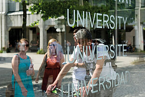 @ Uni Augsburg Transparente Glasscheibe mit den drei Wörtern „University“, „Université“ und „Universidad“ im Vordergrund. Dahinter sind ein Mann auf einem Fahrrad und drei Personen zu sehen, die sich versammelt haben.