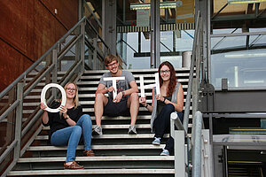 @ OTH Amberg-Weiden Three students sitting on the stairs hold up the letters O, T, and H.