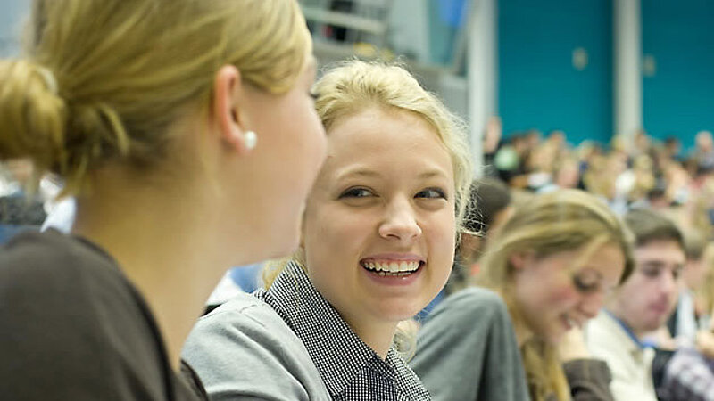 @ Uni Bayreuth A student smiles to her friend while sitting in a lecture hall