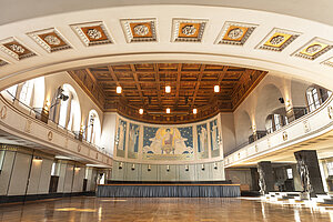 © LMU Das Innere der Hauptaula mit viel Licht. An der Wand befinden sich viele Statuenköpfe und ein großes Wandgemälde.