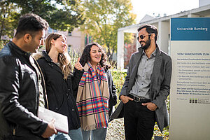 © Benjamin Herges A group of 4 talk and smile together next to a sign that says "Universität Bamberg" and more info