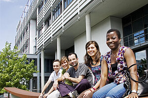 @ Uni Augsburg Studenten verschiedener Ethnien sitzen auf einer Bank und lächeln in die Kamera.