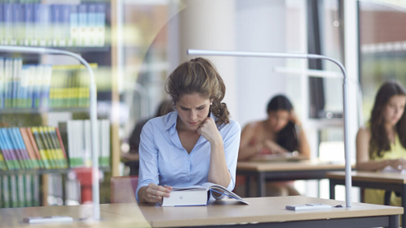 © Jan Greune / LMU Eine Frau sitzt in einer modernen Bibliothek und liest ein dickes Buch. Im Hintergrund lernen andere Studenten.
