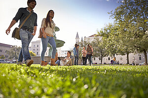 © Jan Greune / LMU Studierende gehen auf dem Rasen vor dem Hauptgebäude der LMU spazieren. Im Hintergrund sitzen einige Studierende auf dem Rasen und unterhalten sich, während eine andere Gruppe im Kreis steht und sich unterhält.