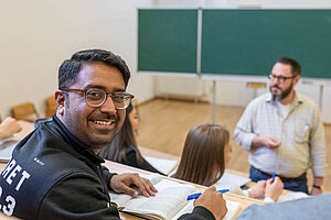 © Benjamin Herges In a lecture hall: a student looks at the camera and smiles, in the background are other students and the lecturer
