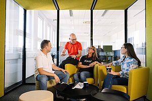 Meeting in a Modern Workspace @Anna Tiessen Four people sitting comfortably in a modern room with yellow furniture, conversating with each other, woman in the middle wears a T-shirt with FAU's logo